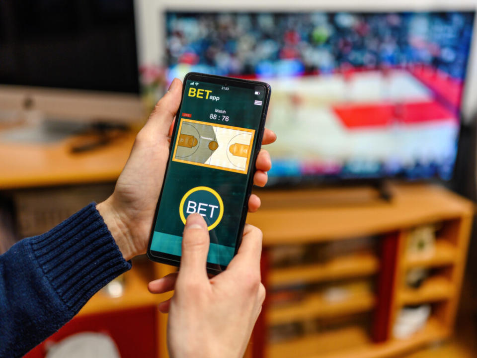 Young man sitting on the sofa at home using a smartphone for sports betting. (Photo: Getty Images/bluecinema)