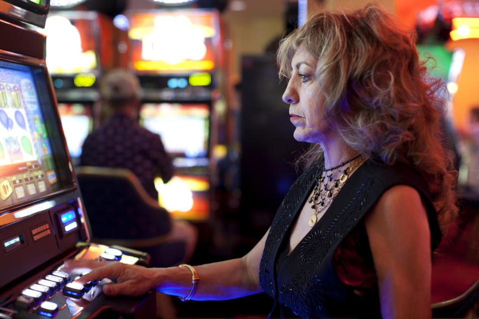 An older woman with curly hair is playing a slot machine in a casino. She is focused on the game. There are other slot machines and a person in the background