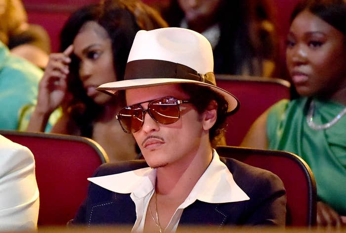 Bruno Mars sits in the audience wearing a fedora, large sunglasses, a wide-collared shirt, and a dark jacket