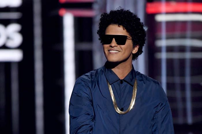 Bruno Mars on stage, wearing sunglasses, a gold chain necklace, and a casual collared shirt