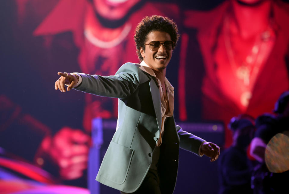 Bruno Mars performs on stage wearing a green blazer, white shirt, and sunglasses. He points towards the audience while smiling