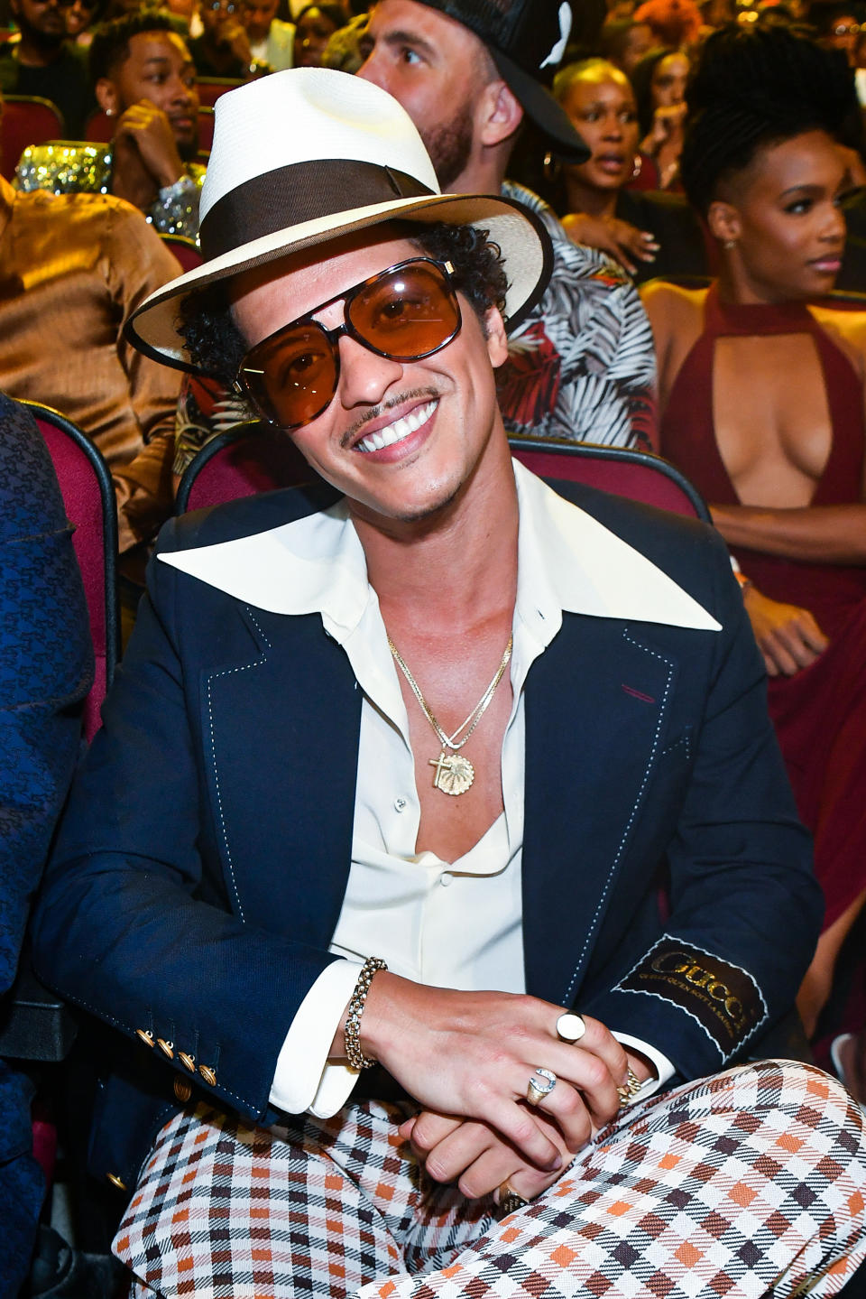 Bruno Mars smiles while sitting, wearing a hat, sunglasses, a dark jacket with a light shirt, checkered pants, and jewelry. Other people are in the background