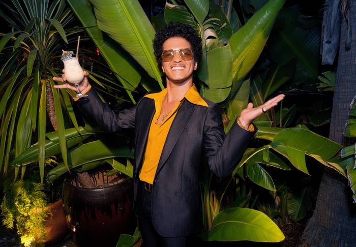 Bruno Mars, wearing a yellow shirt and black suit, holding a cocktail and posing with arms outstretched in a tropical setting