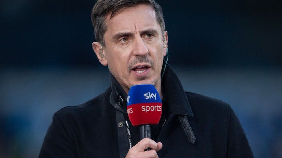 Gary Nevill stands in a football stadium wearing a thick winter coat and holding a microphone, which has a blue and red windshield with Sky Sports' logo printed on it in white. He is also wearing an ear piece made of translucent plastic in each ear, and a clear plastic wire can be seen trailing down into the collar of his coat. He is looking off to the side and appears to be speaking. He's got short black hair and faint stubble on his top lip and chin.