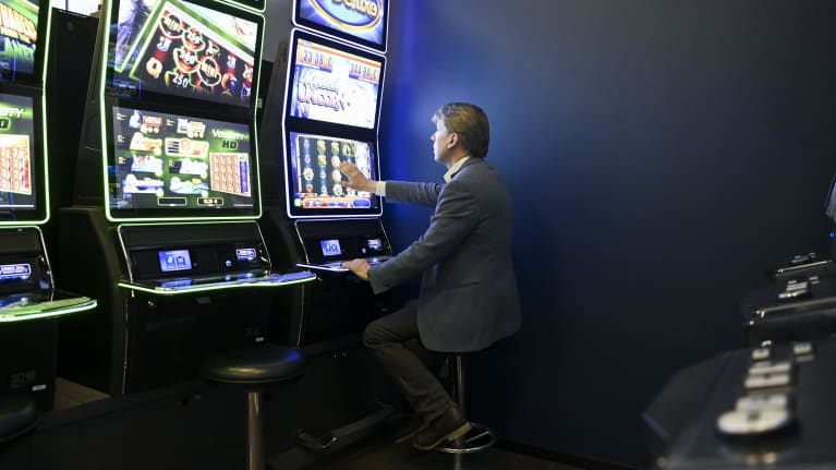 Man wearing a dark grey suit, sitting on a bar stool in front of a brightly coloured slot machine.