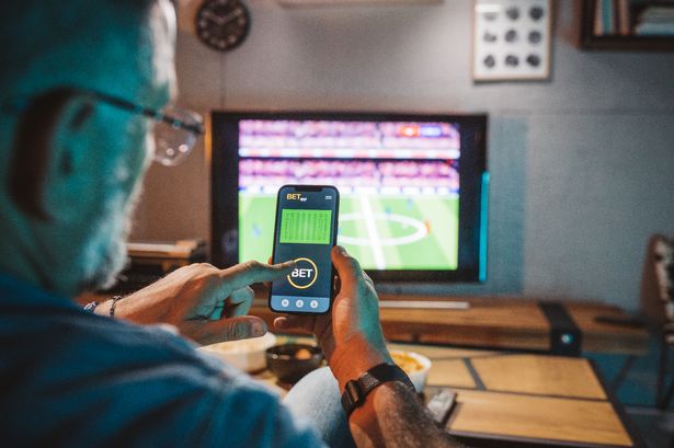 Mature men sitting on sofa at home and watching soccer game.  He is using smart phone for sports betting .