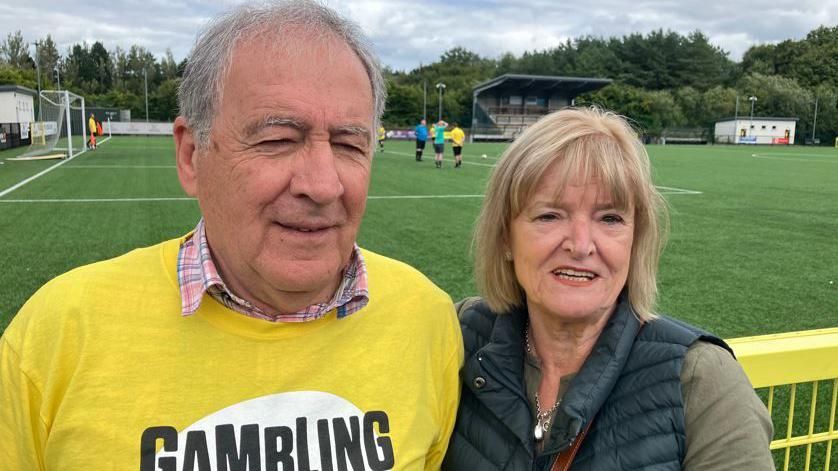 Man and woman standing at the side of a football pitch. He is wearing a yellow jumper, she is wearing a dark gillet.