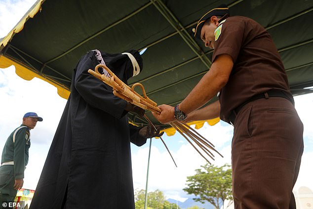 An officer holds a bundle of stiff wooden canes, which he is then seen handing over to an algojo