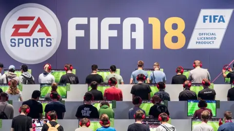 Getty Images Many people are facing away from the camera playing the Fifa 18 video game. Above them, a sign reads Fifa 18.