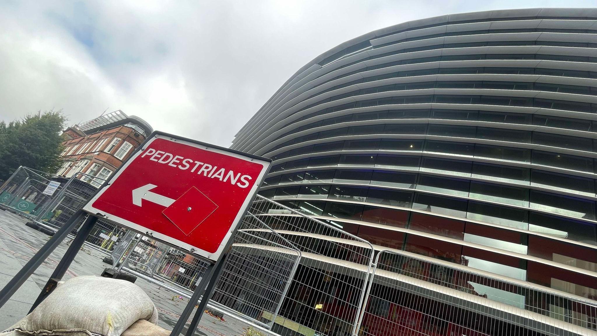 Curve theatre in Leicester with a panel missing from its sunbreaker and fencing in front of it, including a red sign directing pedestrians in front of the building