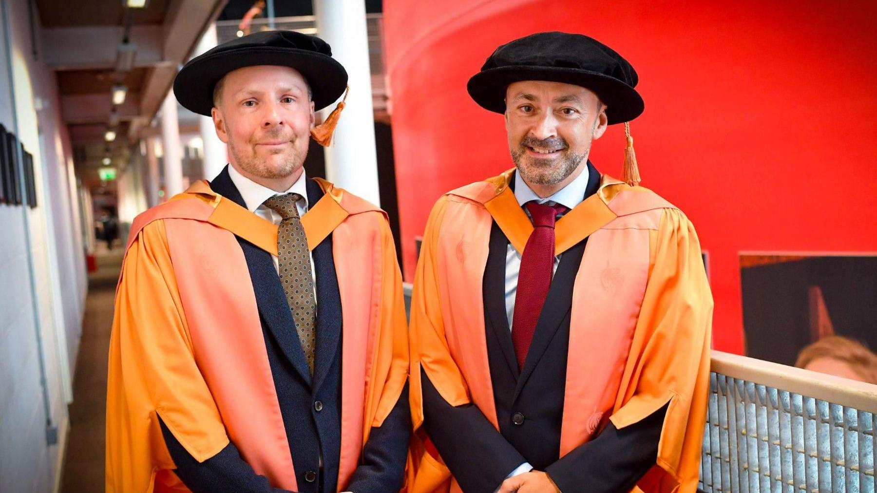 Nikolai Foster pictured on the left and Chris Stafford right. Bot men are wearing orange gowns and black caps. They are also both wearing suits and ties. 