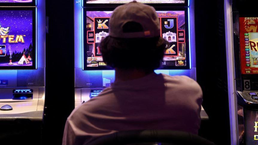 A man wearing a brown baseball cap with his back to the camera, is facing a colourful slot machine