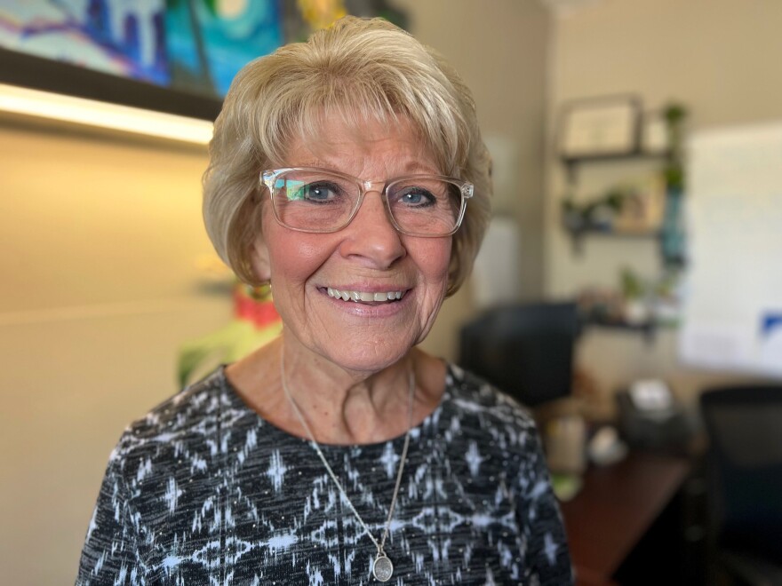 A woman with short blond hair and glasses smiles into the camera. She's standing in a counselor's office.