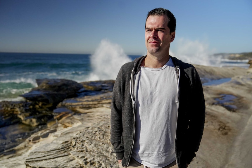 Man with black hair in white t-shirt and grey hoodie stands on beach with waves crashing on rocks behind him on a sunny day