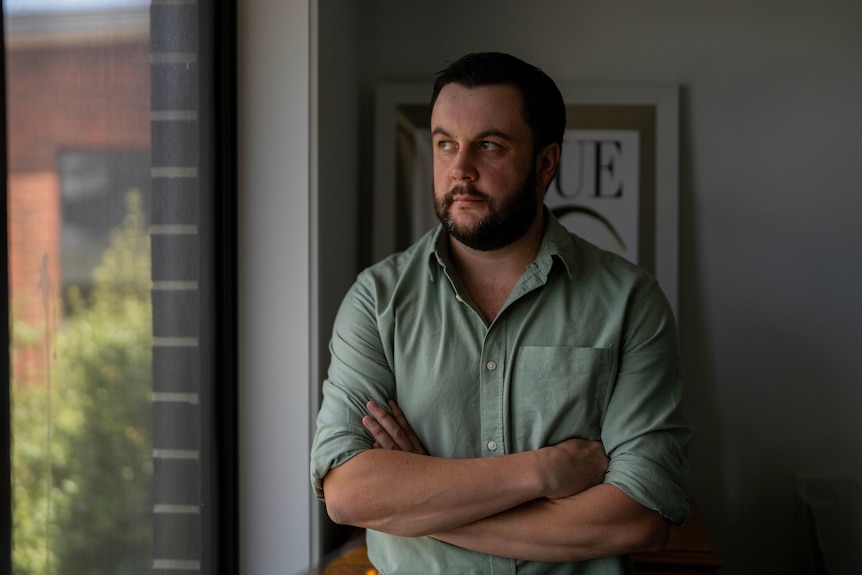 A man stands inside a house, looking out the window with a serious expression.