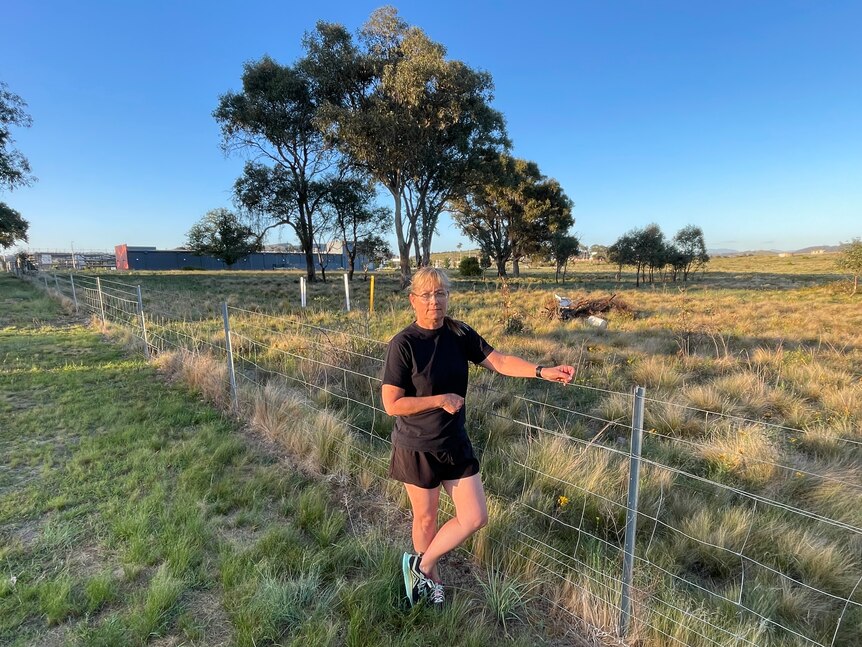 A middle-aged woman wearing a black tee, black shorts, glasses and runners stands by a fence in front of a field.