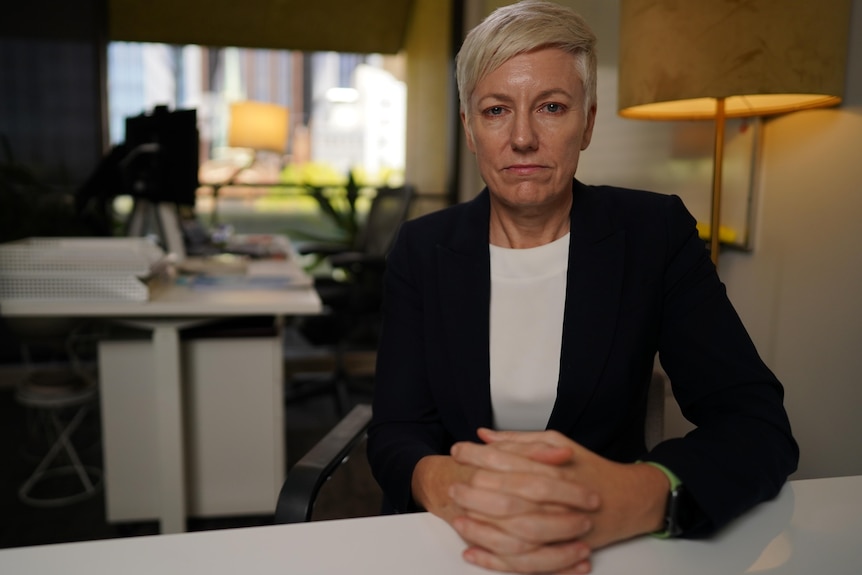 A blonde woman sits at a desk, hands folded.