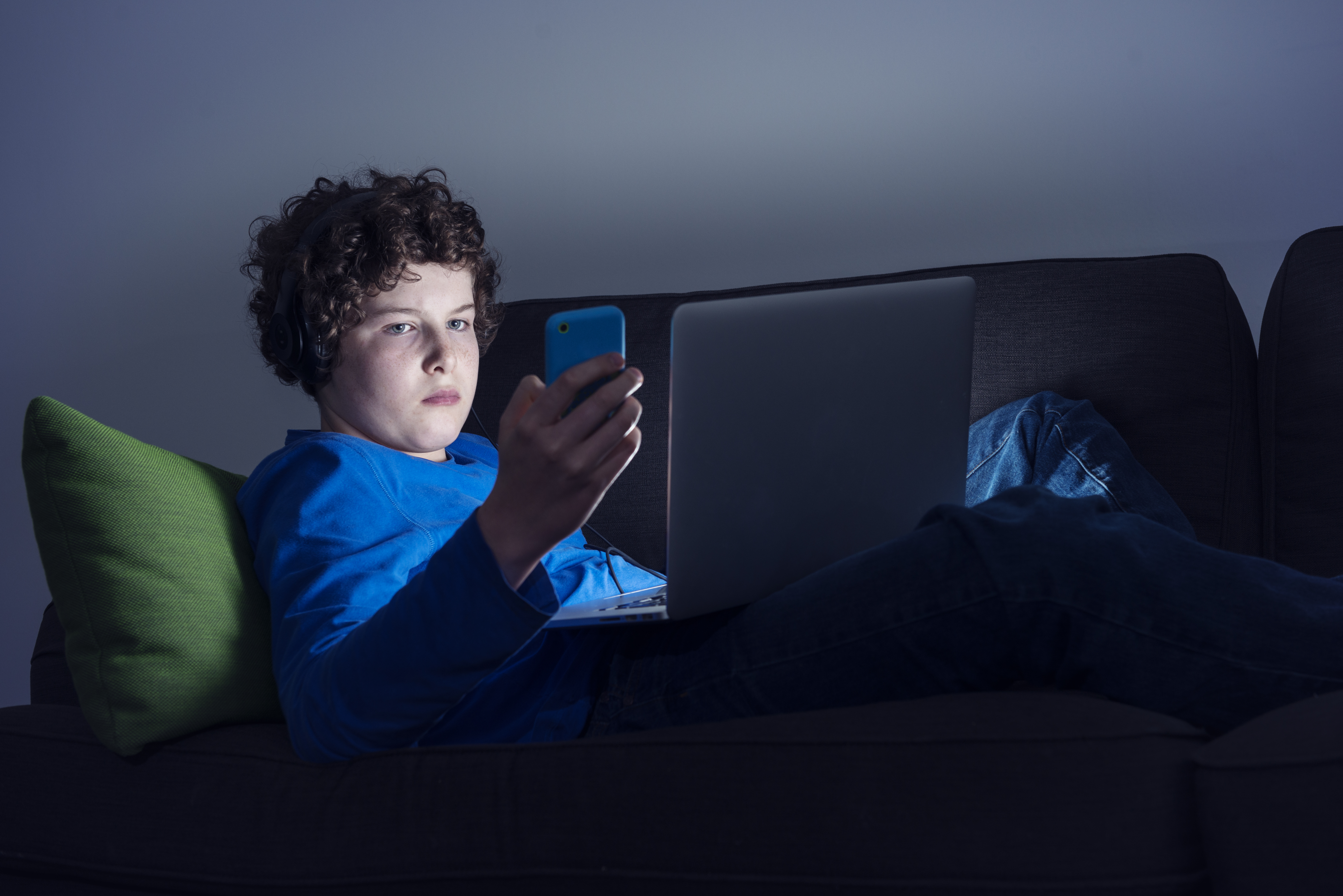 Portrait of a young teenage boy sitting on the sofa at home texting on a smartphone and watching something on screen at the same time. He is illuminated by the light from the screen whilst everything else around him is dark.