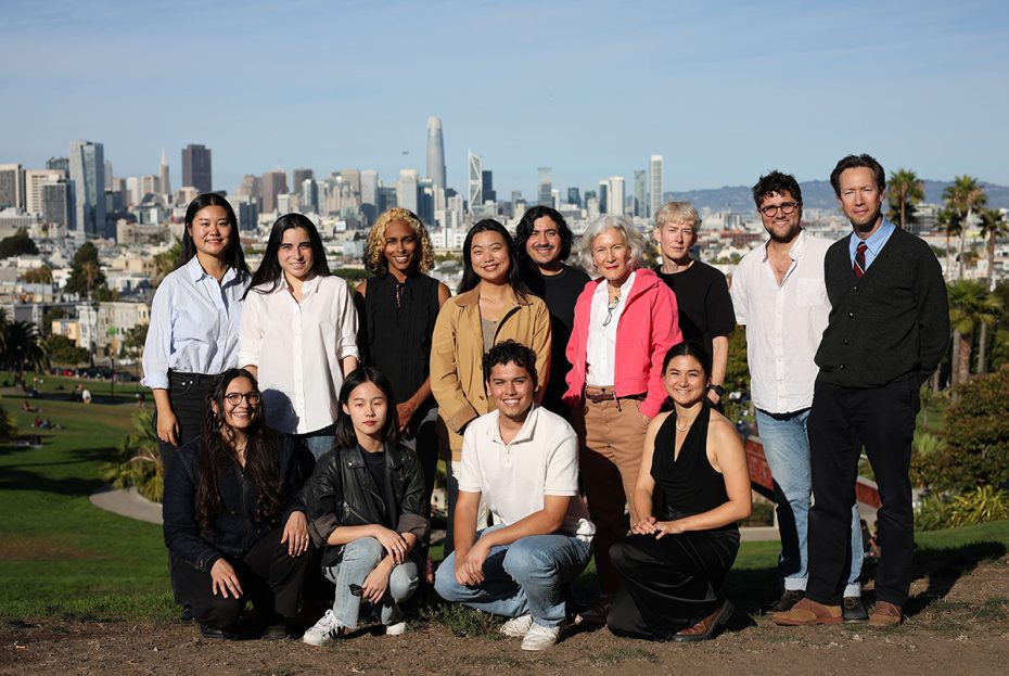 A group of people posing outdoors with a city skyline in the background on a sunny day.